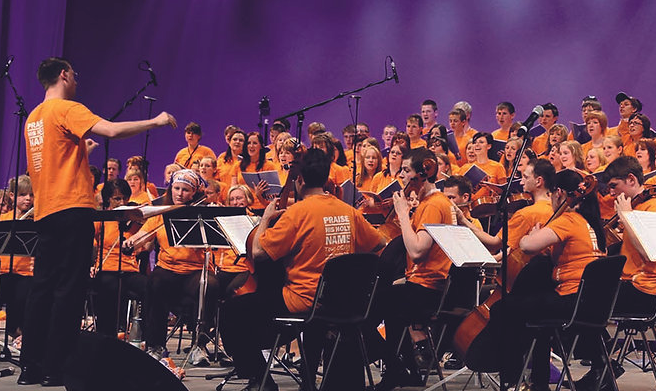 Ein Chor und ein Orchester in orangenen T-Shirts auf einer Bühne